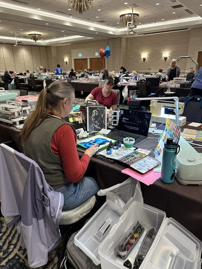A crafter painting at her table, a photo reference propped against her laptop